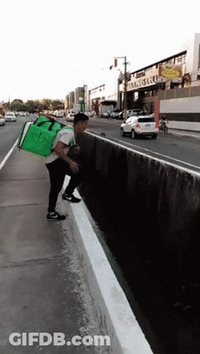 A Man With A Green Bag On His Back Is Standing On A Sidewalk Next To A Street GIF
