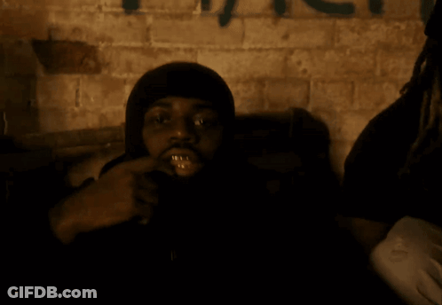 A Man With A Hood On Looks At The Camera With Graffiti On The Wall Behind Him GIF