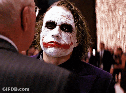 A Man With A Joker Face Painted On His Face Talks To Another Man In A Suit GIF