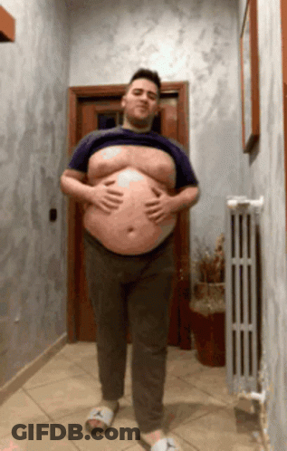A Man With A Large Belly Is Standing In A Hallway Holding His Belly GIF