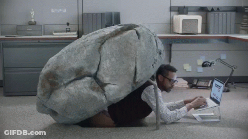 A Man With A Large Rock On His Back Is Using A Laptop In An Office GIF