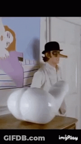 A Man With A Long Nose And A Hat Is Standing Next To A Large White Object On A Table . GIF