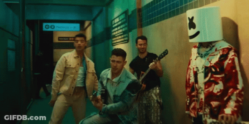 A Man With A Marshmallow Head Stands Next To A Group Of Men Playing Guitars GIF