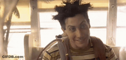 A Man With A Mohawk And A Striped Shirt Is Smiling While Sitting In Front Of A Window . GIF