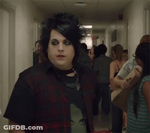 A Man With A Mohawk And Black Eye Makeup Is Standing In A Hallway . GIF