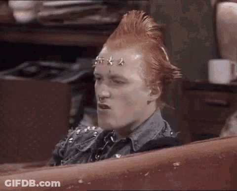 A Man With A Mohawk And Studs On His Head Is Sitting On A Couch And Making A Funny Face . GIF