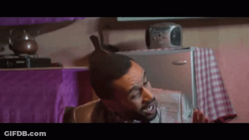 A Man With A Mohawk Is Sitting In Front Of A Refrigerator Holding A Newspaper . GIF