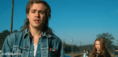 A Man With A Mullet And A Denim Jacket Is Standing In Front Of A Blue Car . GIF