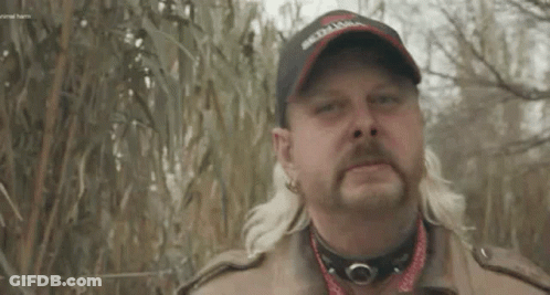 A Man With A Mullet And A Hat Is Standing In Front Of A Fence In A Field . GIF