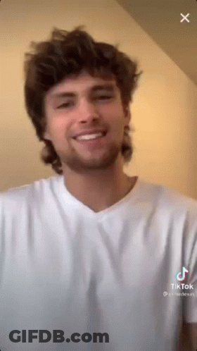 A Man With A Mullet Is Smiling And Wearing A White Shirt . GIF