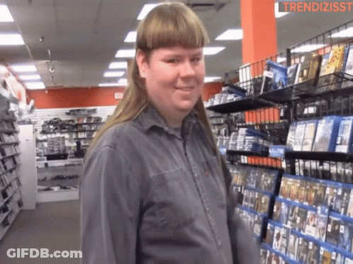 A Man With A Mullet Is Standing In Front Of A Shelf Of Dvds GIF