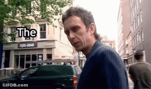 A Man With A Mullet Is Walking Down A Street In Front Of A Building With The Word The On It . GIF
