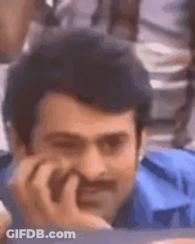 A Man With A Mustache And A Blue Shirt Is Sitting At A Table With His Hand On His Chin . GIF