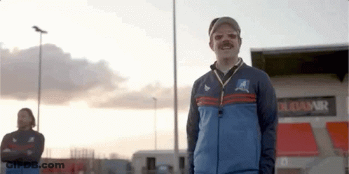 A Man With A Mustache And A Hat Is Standing In Front Of A Stadium . GIF