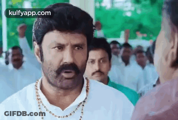 A Man With A Mustache And A Necklace Is Talking To Another Man In A Crowd Of People . GIF