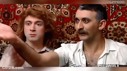 A Man With A Mustache And A Woman With Red Hair Are Sitting Next To Each Other On A Rug GIF