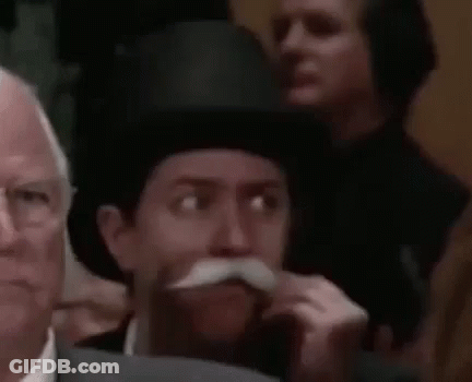 A Man With A Mustache And Top Hat Is Sitting In A Crowd Of People . GIF