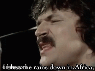 A Man With A Mustache Is Singing Into A Microphone And Saying `` I Bless The Rain Down In Africa '' . GIF