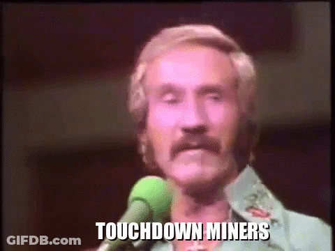 A Man With A Mustache Is Singing Into A Microphone With The Words Touchdown Miners Written On The Screen Behind Him . GIF
