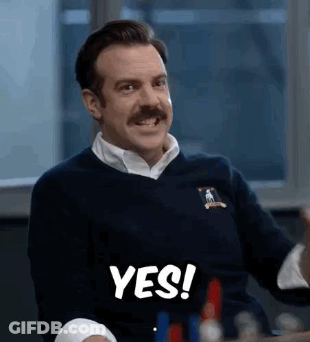 A Man With A Mustache Is Sitting At A Table And Smiling While Holding A Sign That Says Yes . GIF