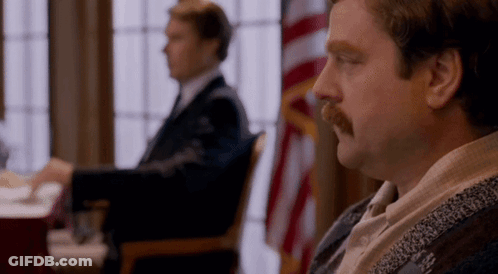 A Man With A Mustache Is Sitting At A Table In Front Of A Flag In A Room . GIF