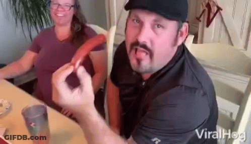 A Man With A Mustache Is Sitting At A Table With A Woman Holding A Hot Dog . GIF