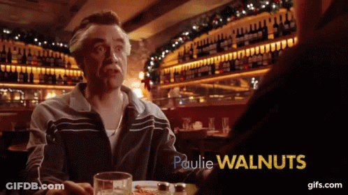 A Man With A Mustache Is Sitting At A Table With Paulie Walnuts In Yellow Letters GIF