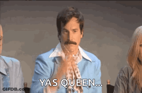 A Man With A Mustache Is Sitting In Front Of A Group Of People And Saying Yas Queen GIF