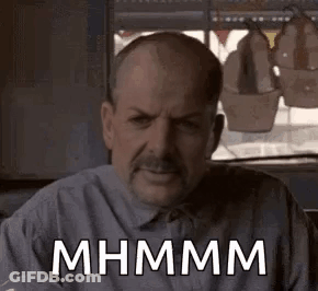 A Man With A Mustache Is Sitting In Front Of A Window With A Sign That Says Mhmm . GIF