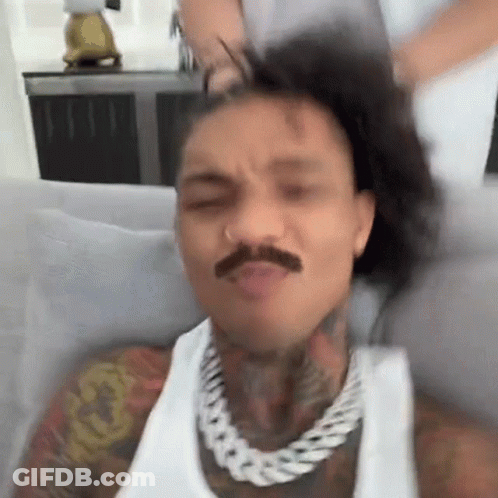 A Man With A Mustache Is Sitting On A Couch With His Hair In A Ponytail . GIF
