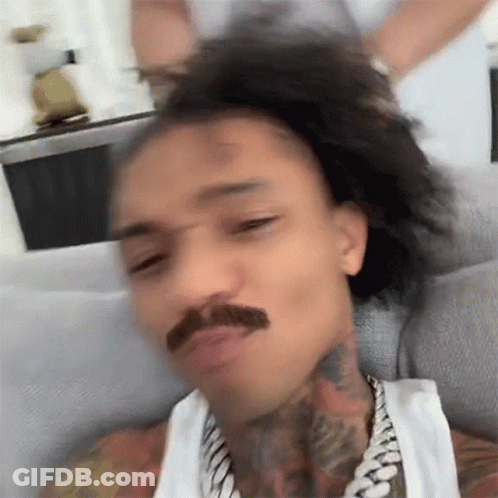 A Man With A Mustache Is Sitting On A Couch With His Hair In A Ponytail . GIF