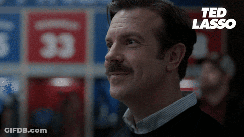 A Man With A Mustache Is Smiling In Front Of A Ted Lasso Sign GIF
