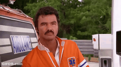 A Man With A Mustache Is Standing In Front Of An Ambulance And Looking At The Camera . GIF