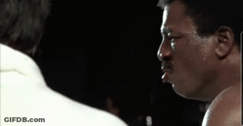A Man With A Mustache Is Standing Next To Another Man In A Boxing Ring In A Dark Room . GIF