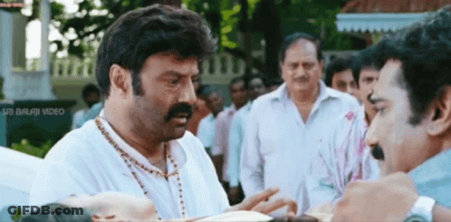 A Man With A Mustache Is Talking To Another Man In Front Of A Sign That Says " Sri Balaji Video " GIF