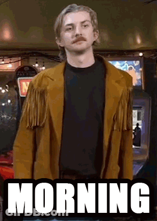 A Man With A Mustache Is Wearing A Brown Jacket And Standing In Front Of An Arcade Machine That Says Morning . GIF
