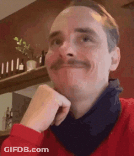 A Man With A Mustache Is Wearing A Red Sweater And A Black Scarf Around His Neck . GIF