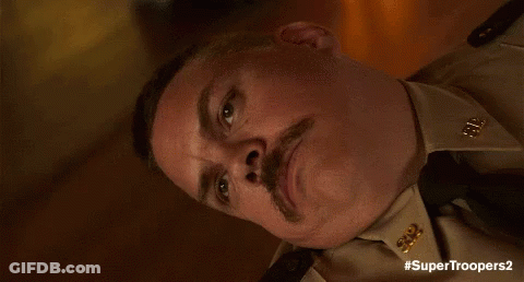 A Man With A Mustache Is Wearing A Tan Uniform And Tie And Has The Hashtag #Supertroopers2 GIF