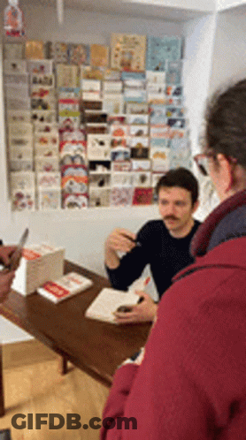 A Man With A Mustache Sits At A Table In Front Of A Wall Full Of Cards GIF