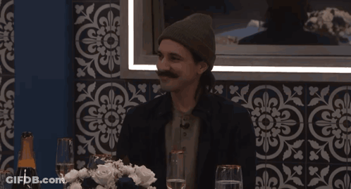 A Man With A Mustache Sits At A Table With Wine Glasses GIF