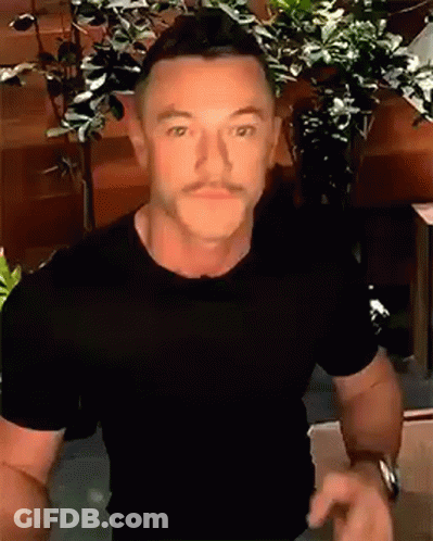 A Man With A Mustache Wearing A Black T-Shirt Is Standing In Front Of A Plant . GIF