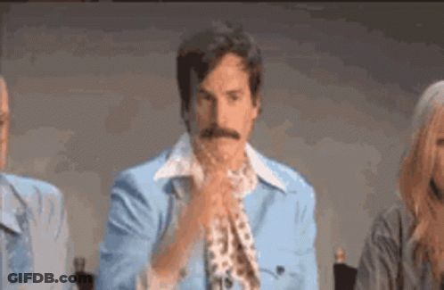 A Man With A Mustache Wearing A Blue Jacket And Tie Is Sitting In Front Of A Microphone . GIF