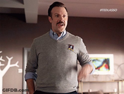 A Man With A Mustache Wearing A Grey Sweater With A Patch That Says " Ted Lasso " GIF