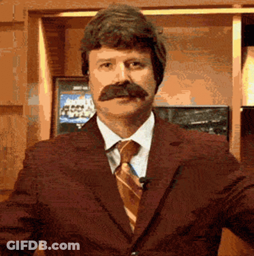 A Man With A Mustache Wearing A Suit And Tie Stands In Front Of A Television GIF