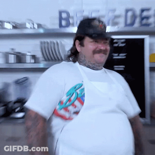 A Man With A Mustache Wearing A White Apron And A Hat Stands In Front Of A Sign That Says Bread GIF