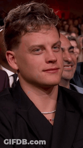 A Man With A Necklace Around His Neck Is Sitting In A Crowd Of People GIF