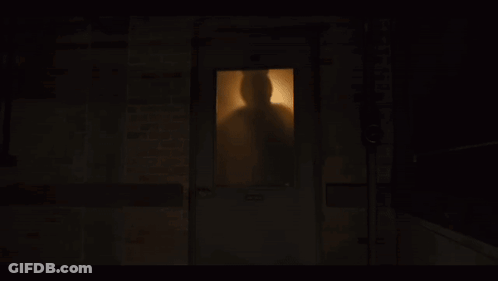 A Man With A Ponytail Is Sitting In A Dark Room Surrounded By Shelves And Boxes GIF