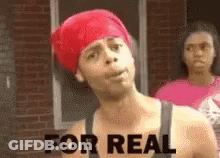 A Man With A Red Bandana On His Head Is Standing In Front Of A Brick Building . GIF