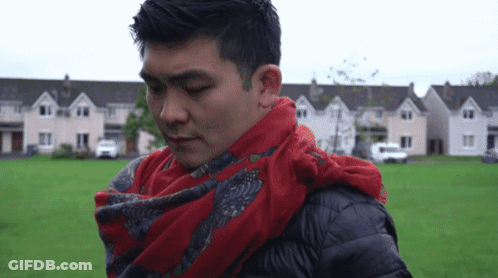A Man With A Red Scarf Around His Neck Is Standing In Front Of A Row Of Houses GIF