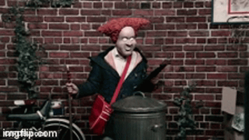 A Man With A Red Wig Is Standing In Front Of A Brick Wall Holding A Trash Can GIF